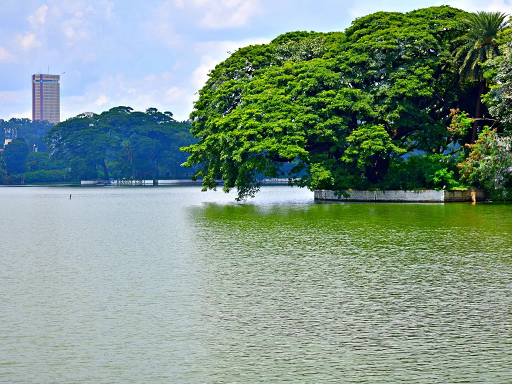 Ulsoor Lake Bangalore partly cloudy day, lush trees lakeside highrise skyline, perfect Karnataka tour package.