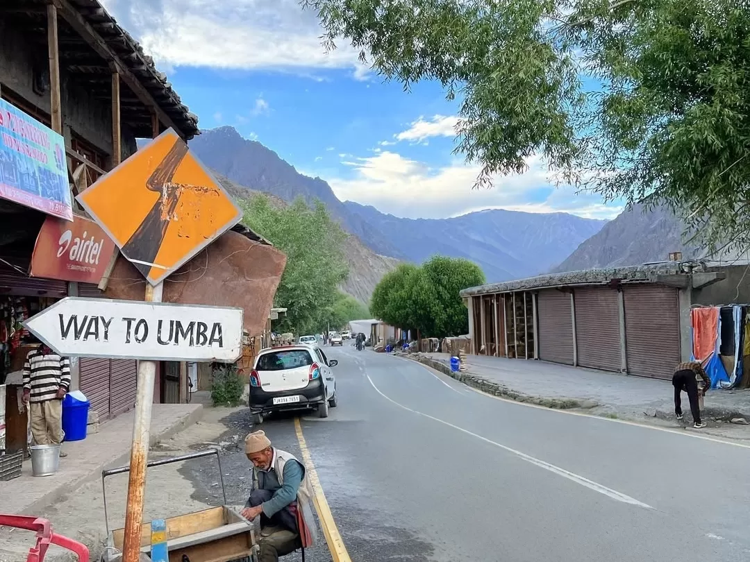 Umba La road sign Kargil Ladakh village street orange Airtel shops shops trees mountains cloudy sky cars local man roadside white SUV gravel road adventure travel photography.