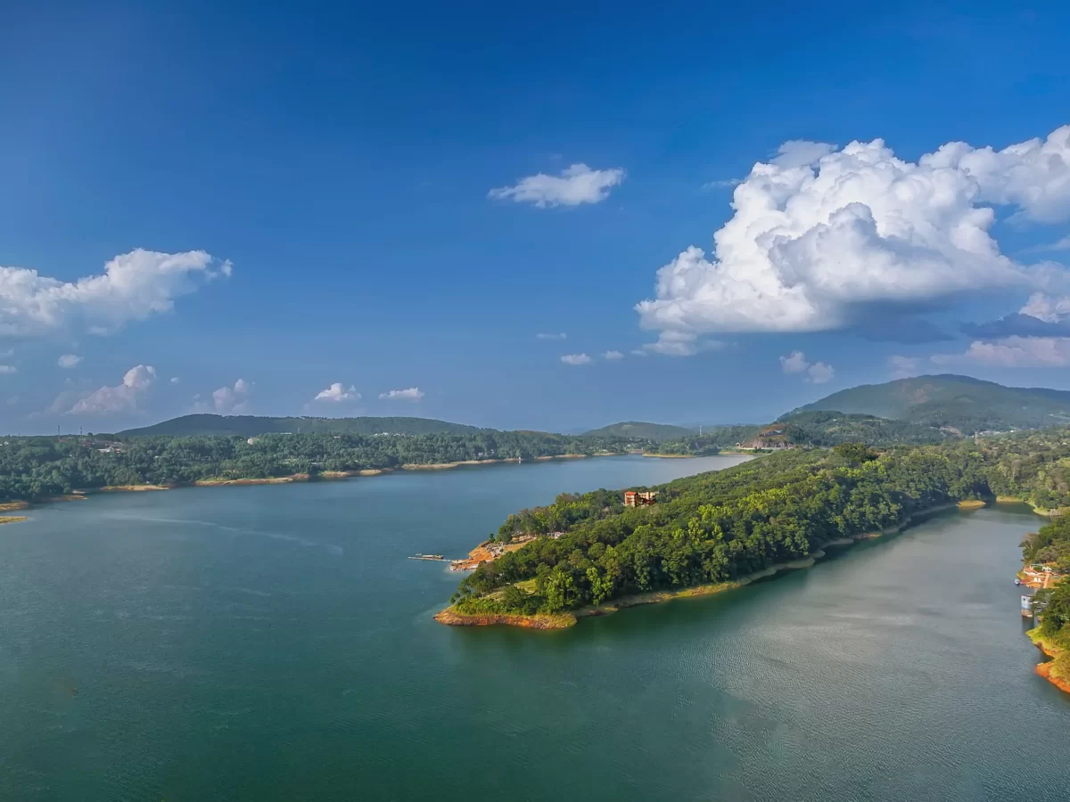 Umiam Lake Shillong Meghalaya aerial panoramic lush green forested peninsula emerald green reservoir distant hills fluffy clouds vibrant blue sky serene hill station landscape, perfect Meghalaya tour packages.