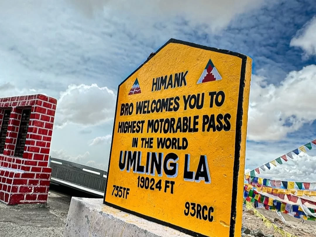 Himank signboard at Umling La Pass Ladakh highest motorable pass during partly cloudy skies, featuring prayer flags and brick wall, perfect adventure Ladakh tour package.