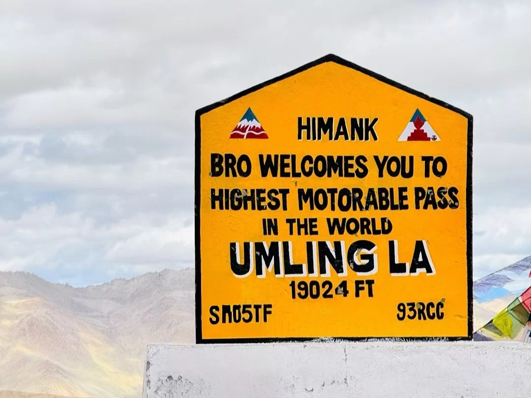 Motivational Himank signboard at Umling La Pass Ladakh during partly cloudy skies, featuring prayer flags and sand dunes, perfect adventure Ladakh tour package.
