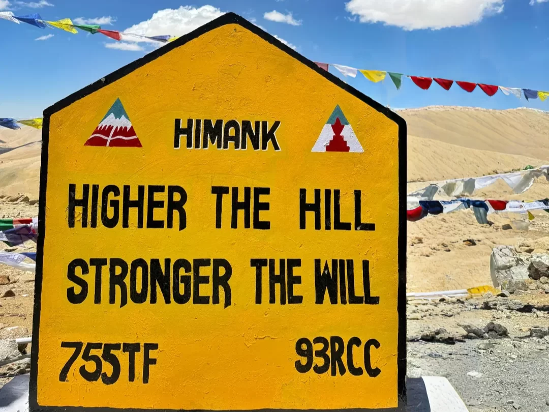 ank motivational signboard at Umling La Pass Ladakh in sunny skies with clouds, featuring prayer flags and desert dunes, perfect adventure Ladakh tour package.