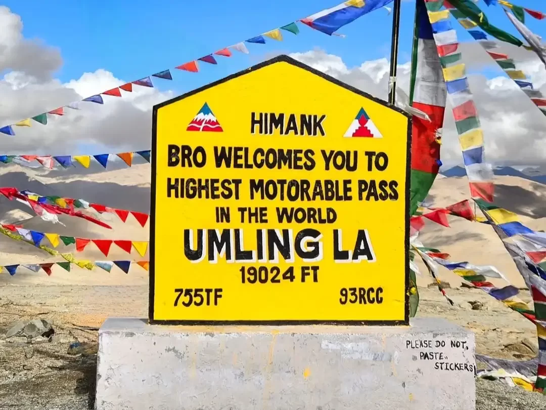 ank signboard at Umling La Pass Ladakh highest motorable pass under partly cloudy skies, featuring colorful prayer flags, perfect adventure Ladakh tour package.