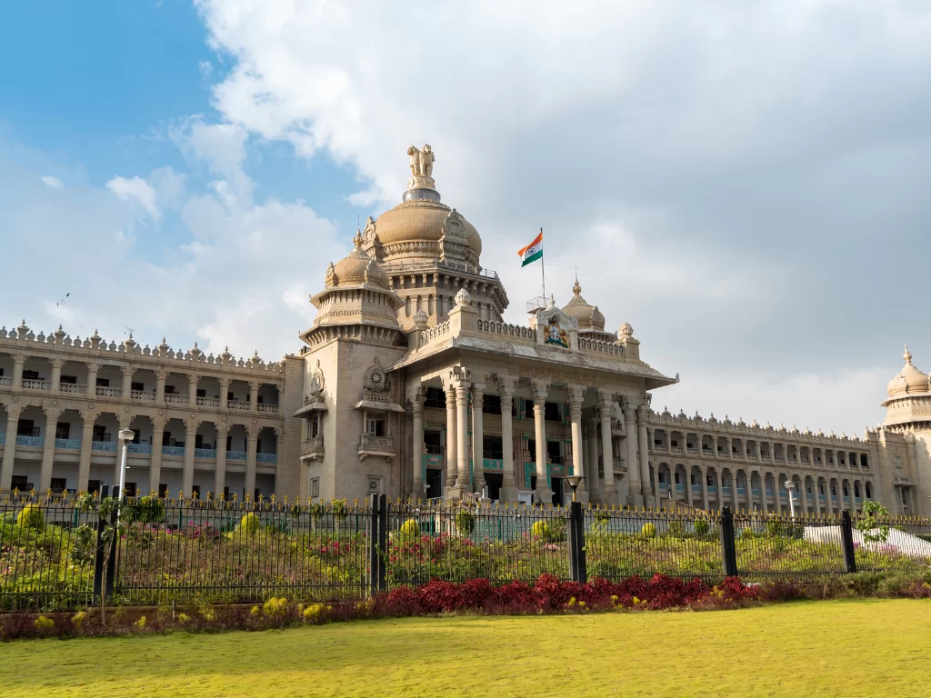 Vidhana Soudha Bangalore partly cloudy sky, golden dome flag pillars arches fences flowers lawn, perfect Karnataka tour package.