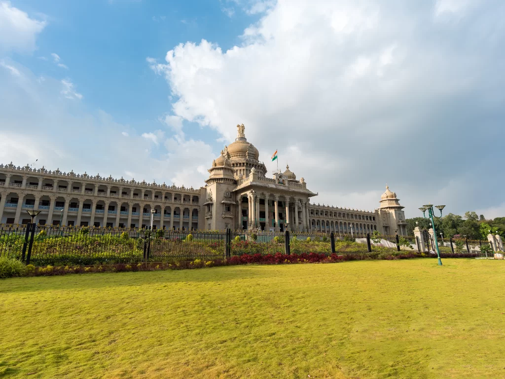 Vidhana Soudha Bangalore partly cloudy day, golden dome flag pillars arches fences flowers lawn lamps, perfect Karnataka tour package.