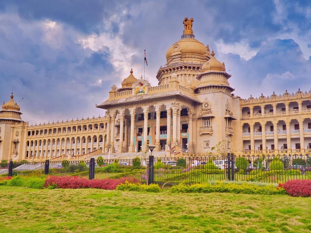 Vidhana Soudha Bangalore cloudy sky, golden dome statue flag pillars domes arches fences flowers cars grass, perfect Karnataka tour package.