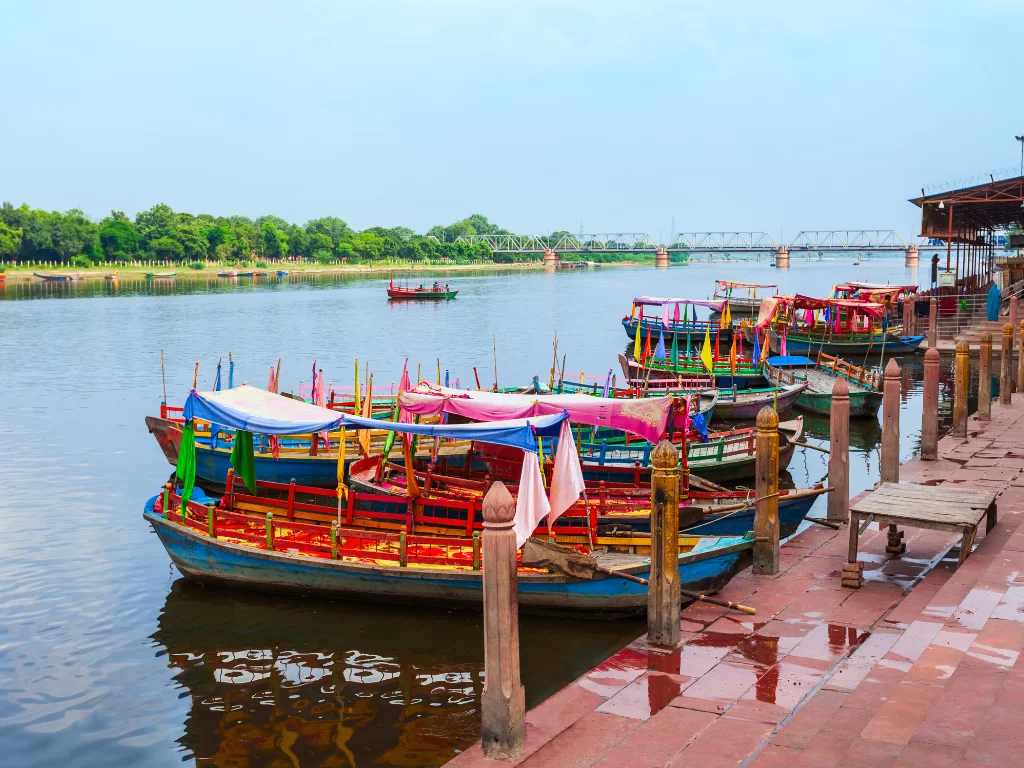 Colorful flower boats docked at Vishram Ghat Mathura on Yamuna River in clear weather, featuring vibrant hulls and red sandstone steps, perfect cultural Mathura Vrindavan tour package.