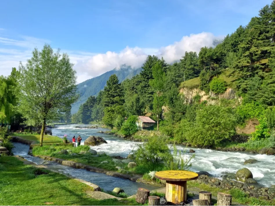Tourists by Lidder River at Aru Valley Pahalgam during sunny day, featuring pine forests meadows cottages and mountains, perfect adventure experience Kashmir tour packages.