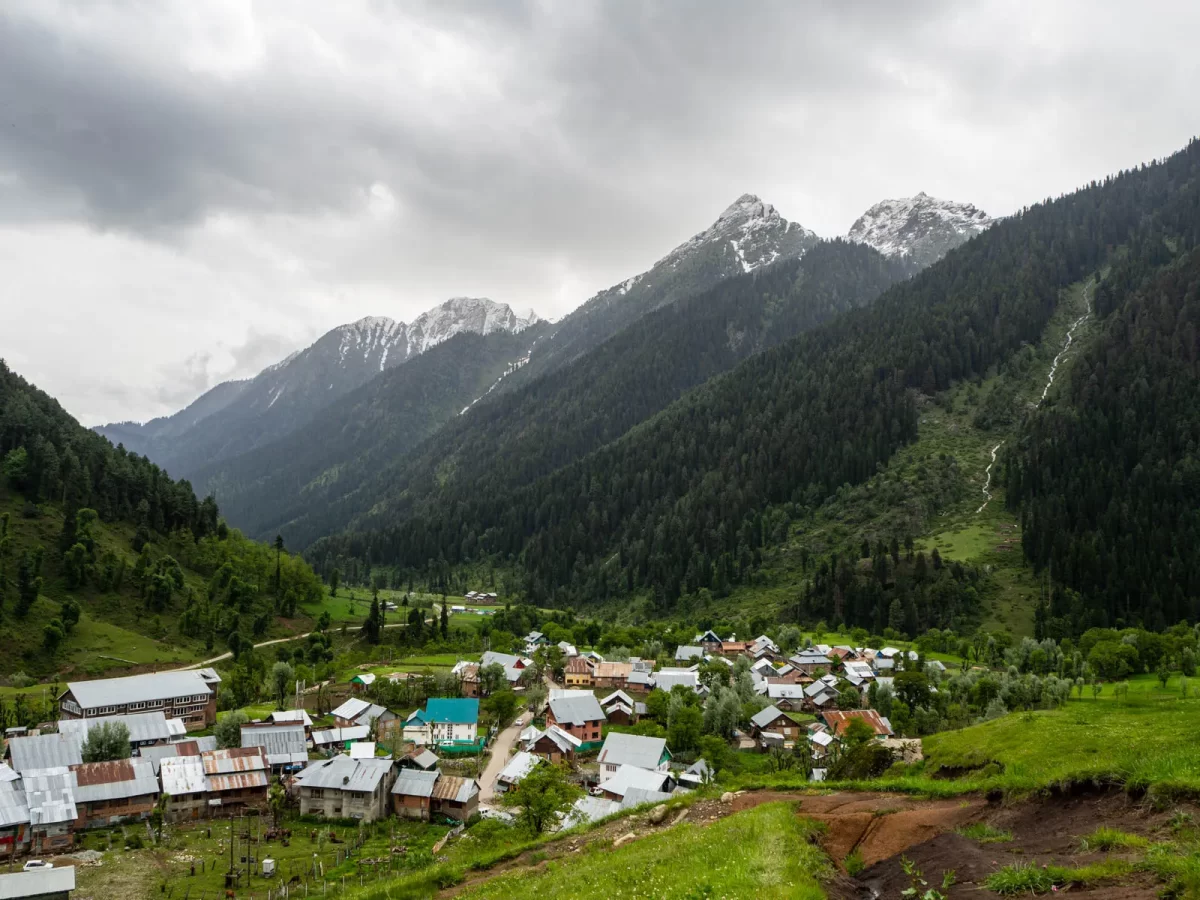 Traditional village houses in Aru Valley Pahalgam during cloudy day, featuring pine forests green meadows and snow peaks, perfect adventure experience Kashmir tour packages.