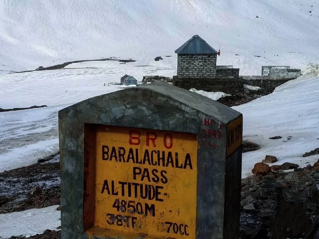 Baralachala Pass signboard at 4850m with stone hut amid snowy peaks during clear day, featuring BRO emblem and rugged terrain, perfect adventure Manali-Leh highway Ladakh tour package.
