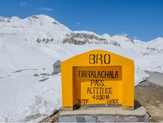 BRO signboard at Baralachala Pass 4850m during sunny skies, featuring yellow plaque and snow peaks, perfect high-altitude adventure Ladakh tour package.