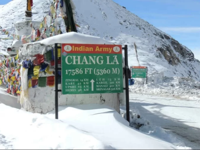 Indian Army sign at Changla Pass 17586 ft (5360m) during clear skies, featuring prayer flags, Leh routes, snowy backdrop, perfect adventure Ladakh tour package