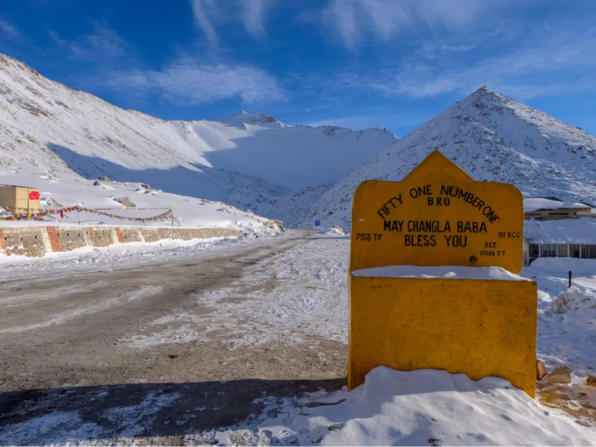 May Changla Baba Bless You BRO signboard at Changla Pass during clear skies, featuring snowy Himalayan valley and road, perfect spiritual adventure Ladakh tour package.