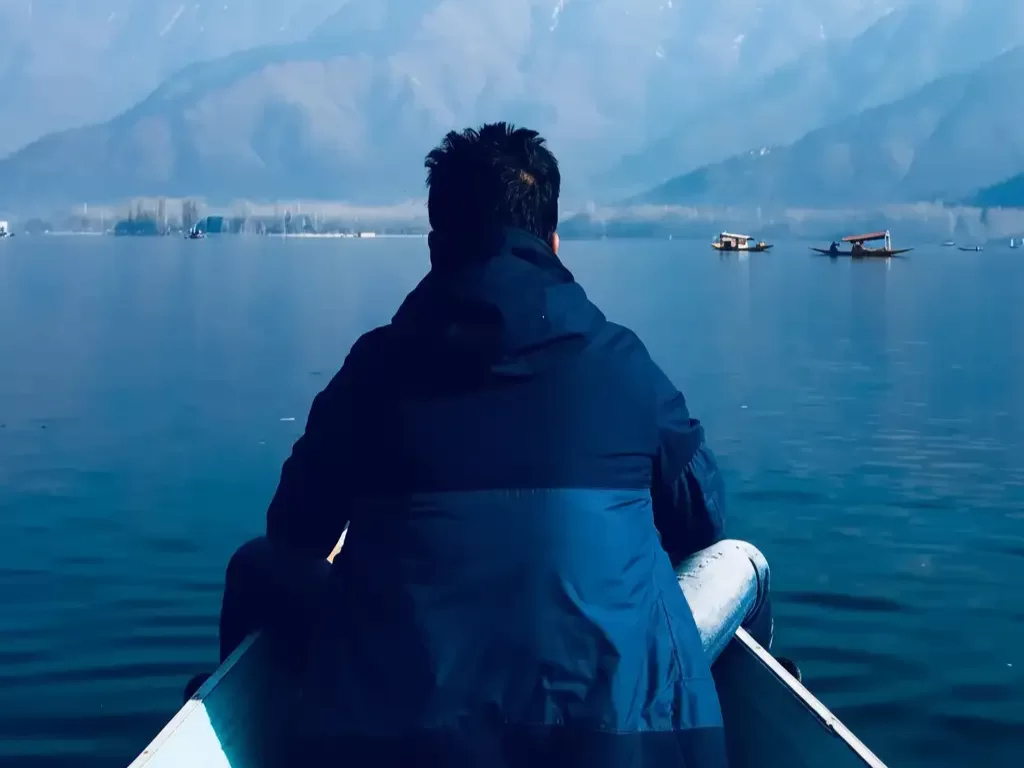 Back view tourist shikara boat Dal Lake Srinagar snowy mountains distant shikaras misty reflection featured in Kashmir Srinagar tour packages 