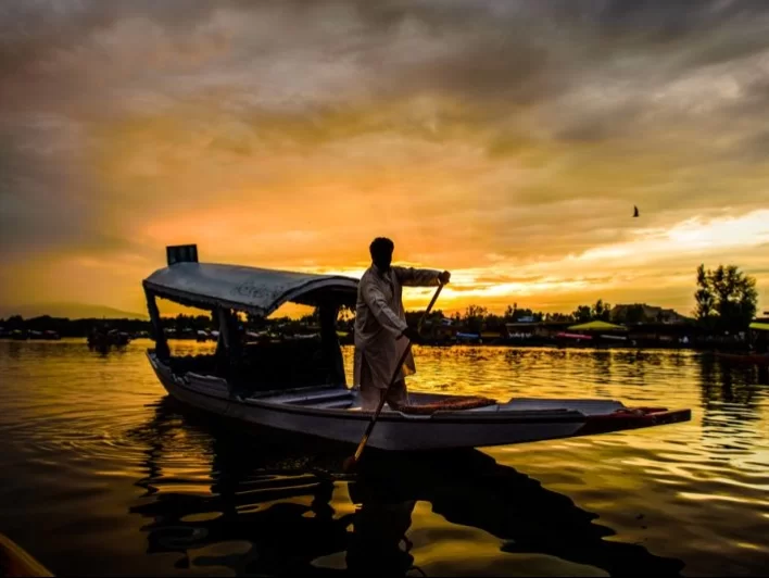 Shikara rower silhouette sunset Dal Lake Srinagar with houseboats orange sky reflection featured in Kashmir Srinagar shikara tour packages 