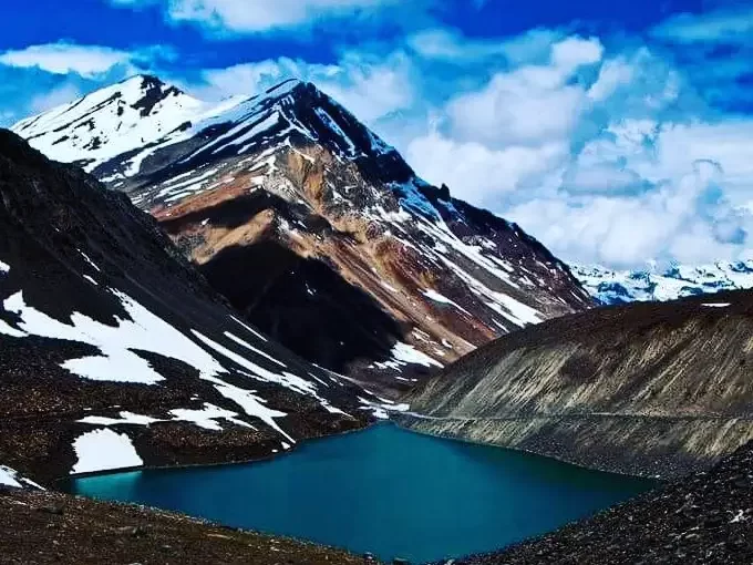 Deepak Tal Lahaul Spiti Himachal Pradesh India vibrant turquoise glacial lake nestled dramatic snow capped peaks rugged brown mountains blue skies scattered clouds foreground rocks highway edge, perfect Manali Leh highway scenic tour package