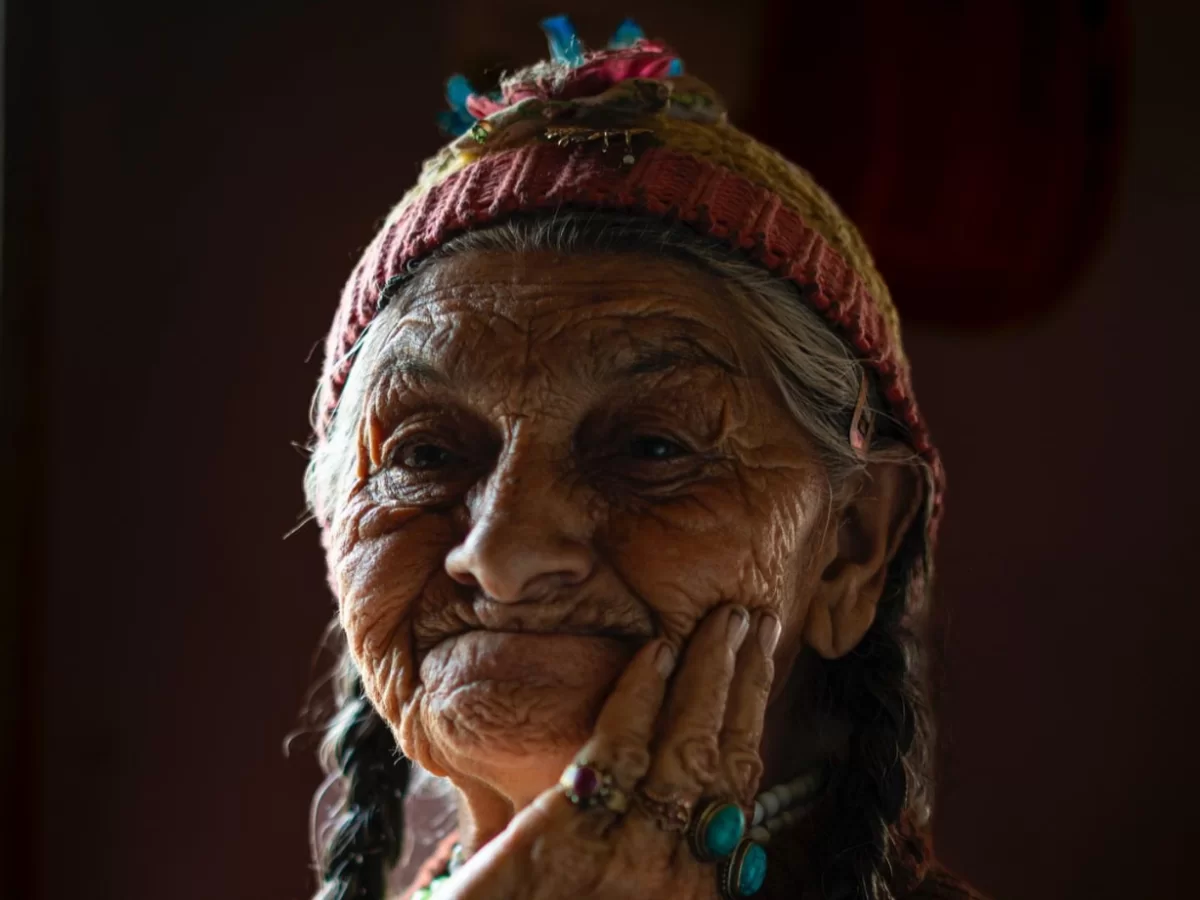 Elderly Dha Hanu woman during soft light, featuring traditional red hat, turquoise jewelry, and braided hair, perfect cultural experience for Ladakh Dha Hanu tour package. 