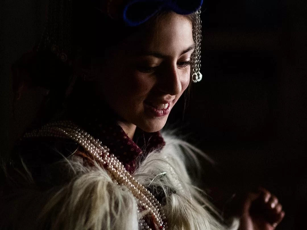 Dha Hanu woman during dramatic light, featuring blue headdress with beads, pearl necklaces, and fur-trimmed attire, perfect cultural experience for Ladakh Dha Hanu tour package. 