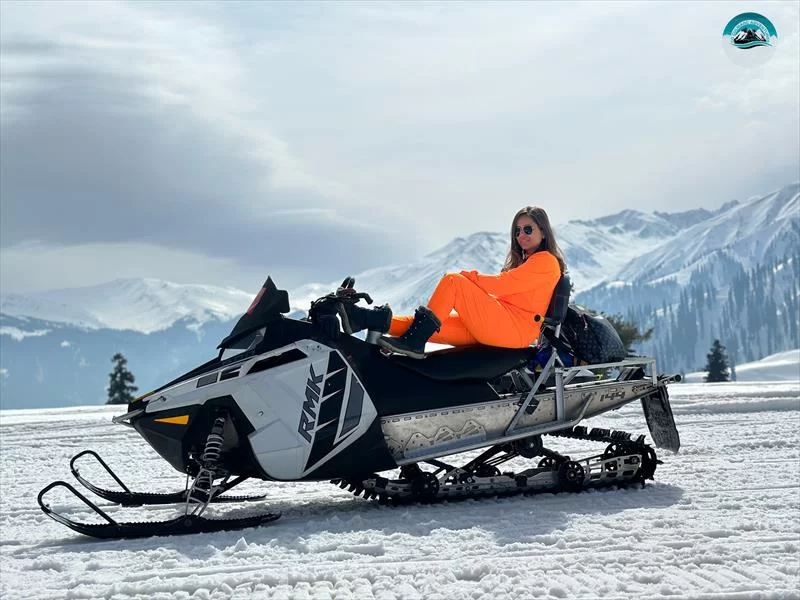Gulmarg Kashmir snow slope orange suit woman black white snowmobile sunglasses snowy mountains cloudy skies pine trees, perfect winter snowmobile adventure tour package. 