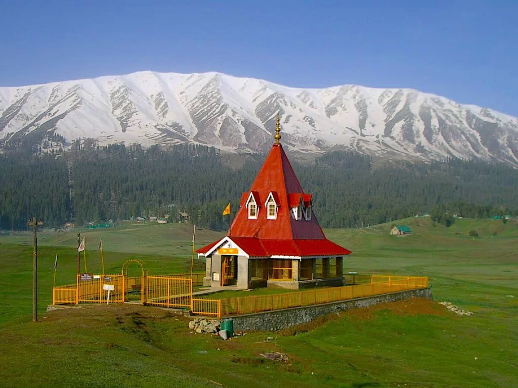 Gulmarg Kashmir red temple Shiva Mahadev shrine snow capped mountains green meadows yellow fence flags blue skies, perfect Himalayan spiritual pilgrimage tour package. (