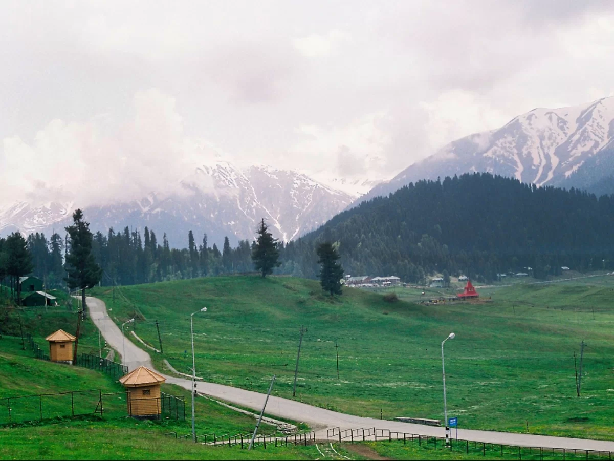 Gulmarg Kashmir green meadows winding road wooden huts red temple snowy mountains pine forests cloudy skies lamp posts, perfect scenic highland meadow tour package.