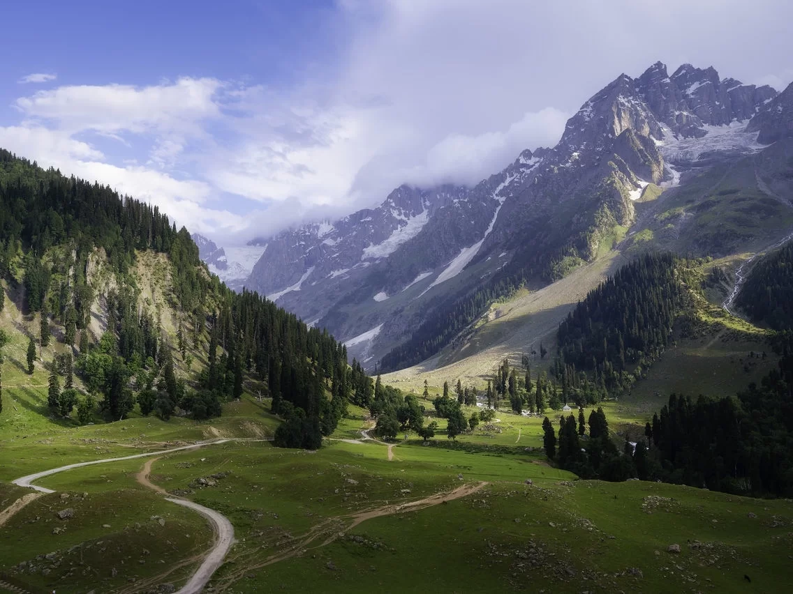 Gulmarg Kashmir Apharwat peak snowy mountains pine forests green meadows winding paths huts clouds blue skies, perfect gondola valley panoramic Himalayan adventure tour package.