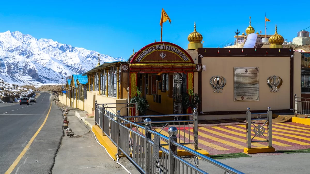 Gurdwara Shri Pathar Sahib Leh Ladakh with golden domes saffron flag intricate signage roadside setting against snow-capped Himalayan peaks blue skies vehicles and railings, sacred Sikh heritage adventure Ladakh tour package highlight.
