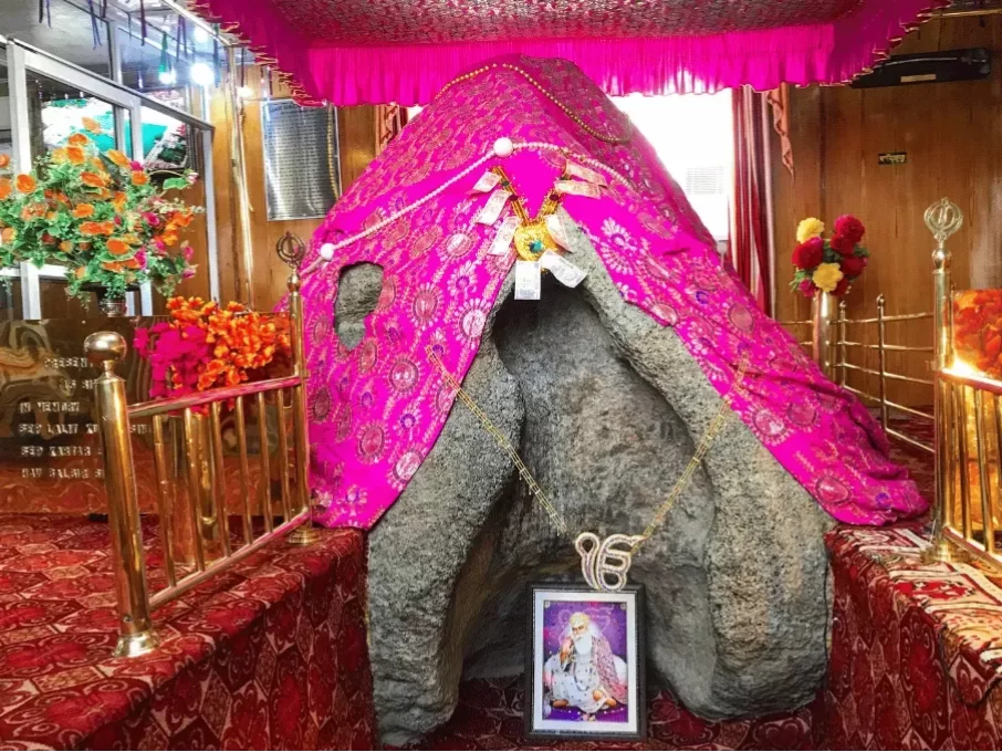 Sacred Pathar Sahib boulder draped in vibrant pink canopy with Guru Gobind Singh portrait floral tributes golden railings ornate carpet lighting and framed Sikh emblem at Gurdwara Pathar Sahib Leh Ladakh, iconic miracle rock spiritual Ladakh tour package 