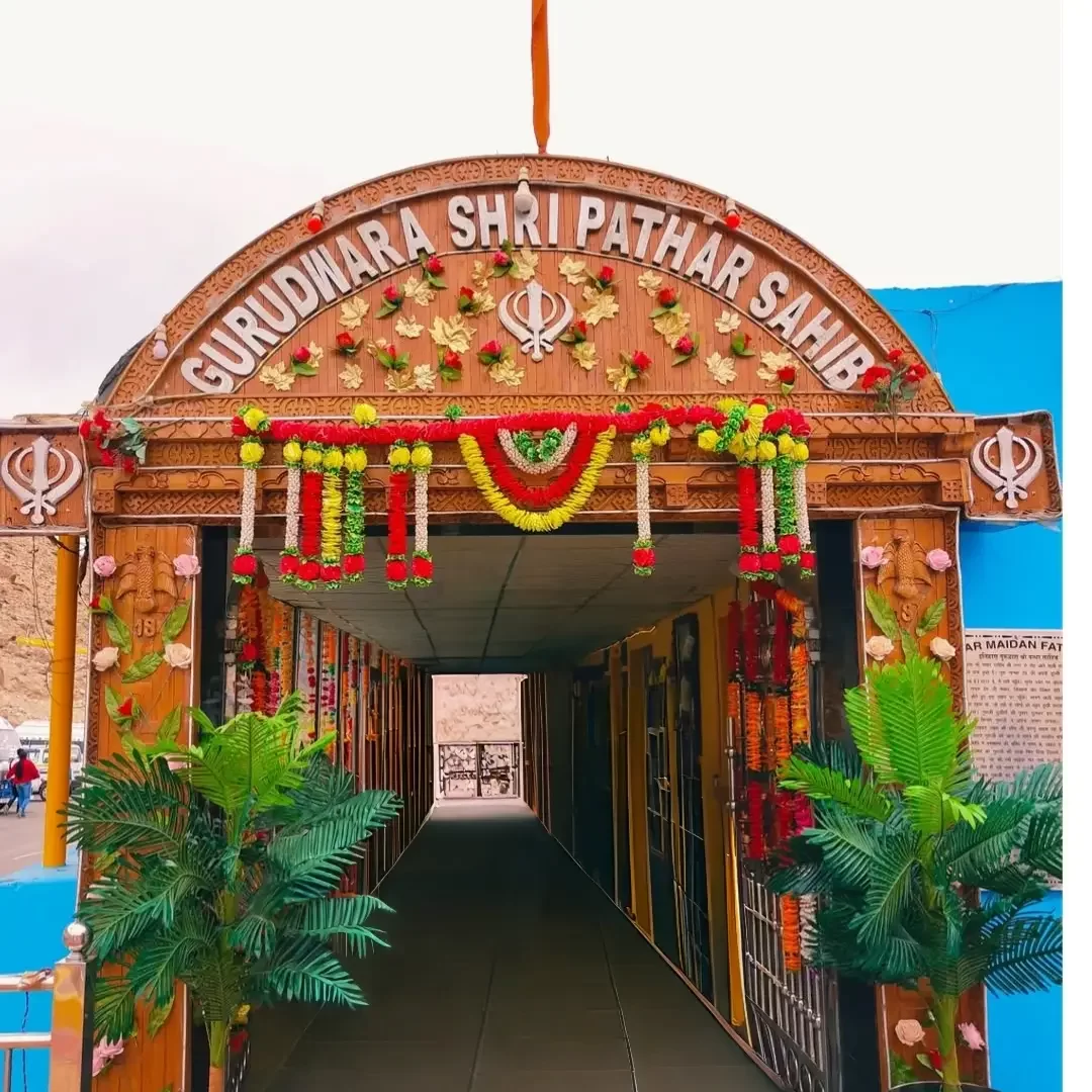 Ornate duplicate entrance archway at Gurdwara Shri Pathar Sahib Leh Ladakh showcasing carved wooden gateway saffron flag floral marigold garlands Khanda emblems green plants corridor blue walls rocky terrain vehicles, colorful inviting spiritual Ladakh to