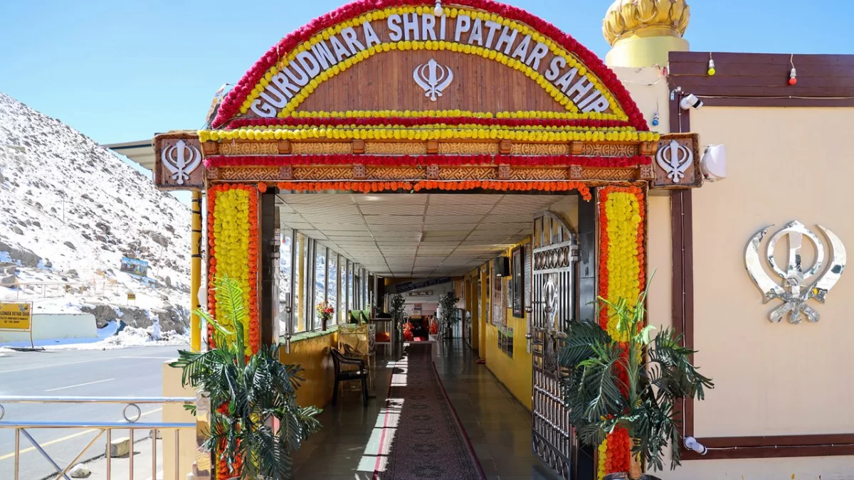 Vibrant floral-adorned wooden gateway entrance at Gurdwara Shri Pathar Sahib Leh Ladakh with marigold yellow garlands Khanda symbols saffron flag dome golden dome potted plants corridor snowy mountains roadside sign, festive spiritual Ladakh tour package 