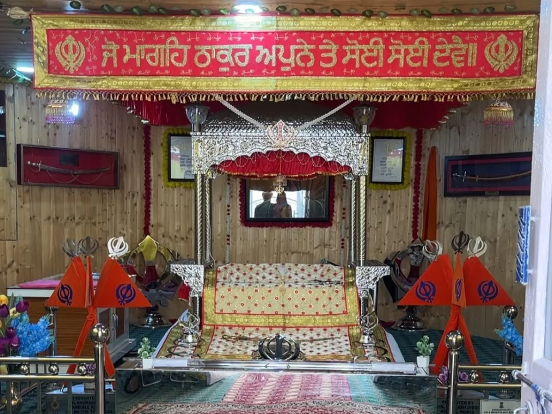 Guru Granth Sahib swing palanquin shrine at Gurdwara Shri Pathar Sahib Leh Ladakh featuring red embroidered canopy Punjabi Mata Gujri Nagar Kaur banner Khanda symbols floral pink stands wooden walls Kirpans portraits, sacred devotional Sikh palanquin Lada