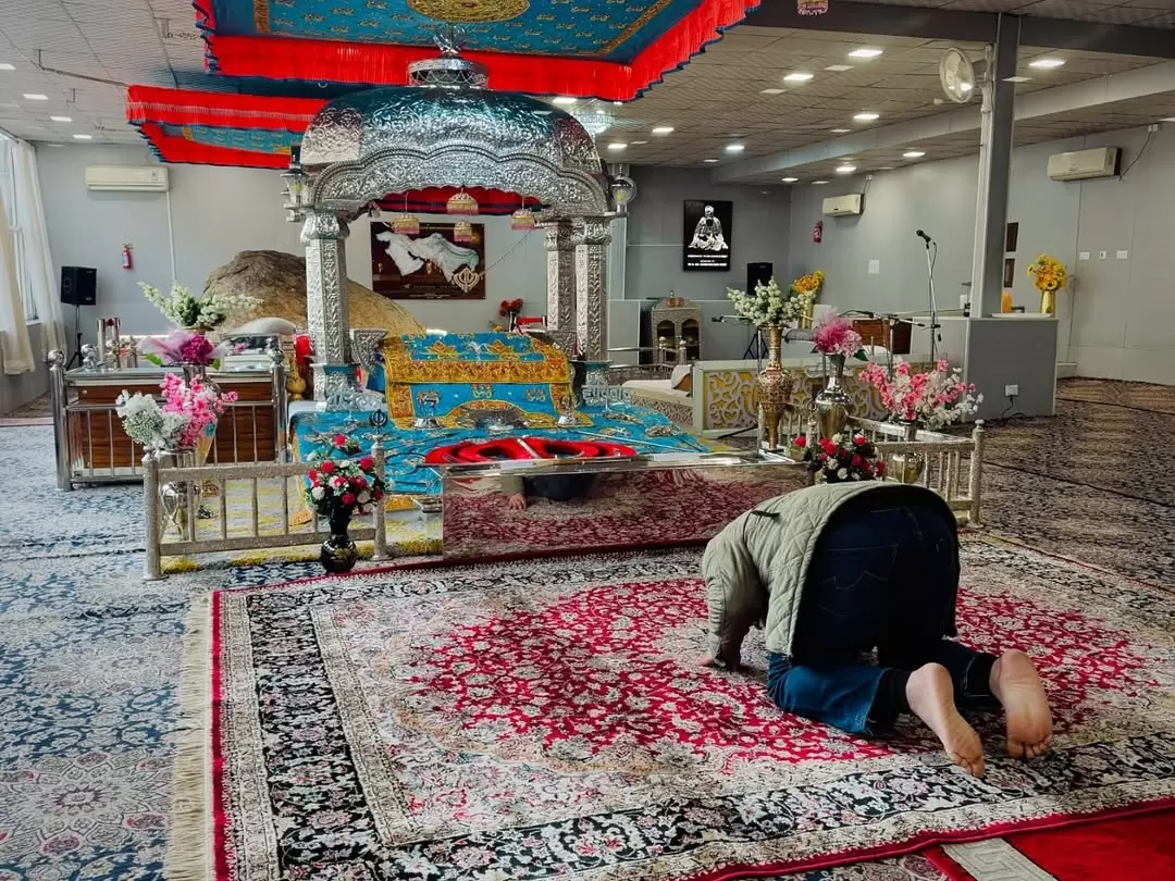 Main prayer sanctum at Gurdwara Shri Pathar Sahib Leh Ladakh with elevated golden Guru Granth Sahib canopy red floral hangings Khanda symbols potted flowers devotee prostrating red carpet sofas rock backdrop AC units, reverent worship space Ladakh tour pa