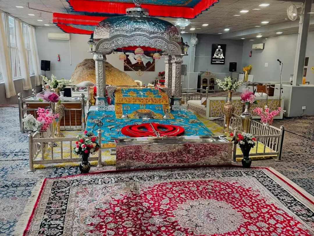 Alternate view main prayer sanctum Gurdwara Shri Pathar Sahib Leh Ladakh showcasing golden domed Guru Granth Sahib canopy red hangings blue platform floral vases Sacred Pathar backdrop sofas speakers AC units intricate carpets, holy worship area Ladakh to