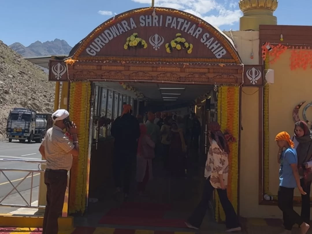 Busy main entrance Gurdwara Shri Pathar Sahib Leh Ladakh with carved archway saffron flag Khanda symbols garlands diverse pilgrims headscarves entering corridor bus parked Himalayan mountains highway roadside, lively spiritual hub Ladakh tour package attr