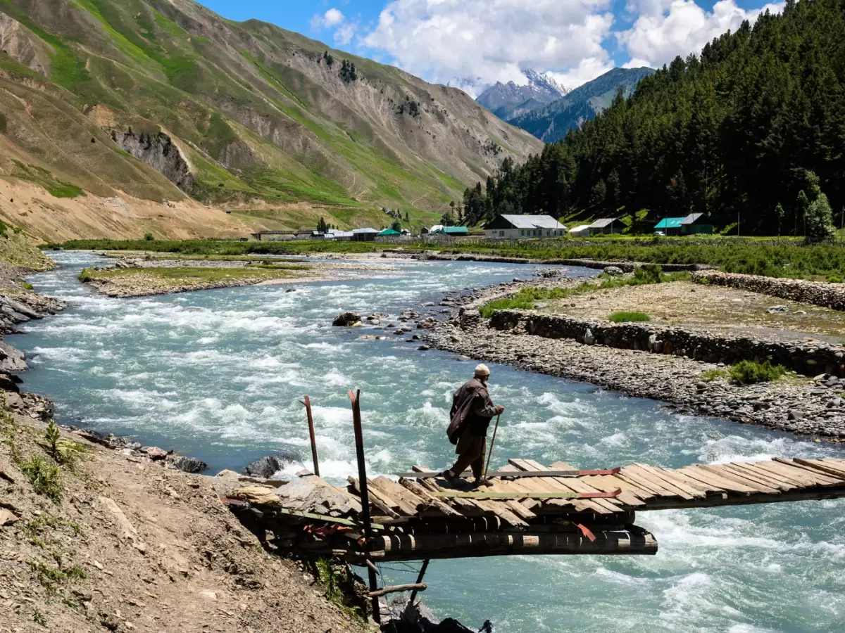 Elder crossing wooden bridge at Gurez Valley during sunny partly cloudy weather, featuring turquoise river and green mountains, perfect adventure Srinagar tour package. 
