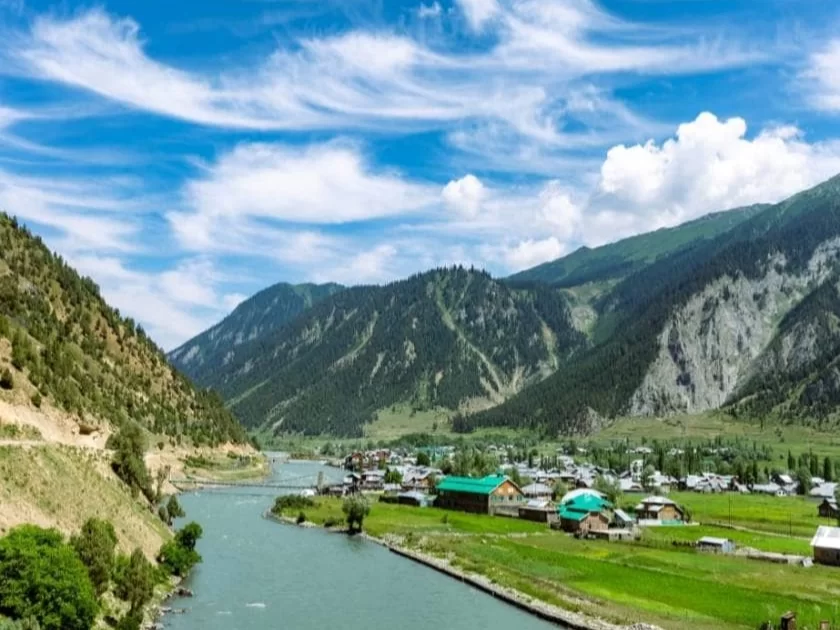 Gurez Valley riverside Kashmir sunny day, featuring green roofed villages turquoise river lush green fields forested mountains blue skies clouds, perfect cultural adventure Srinagar Kashmir tour packages.