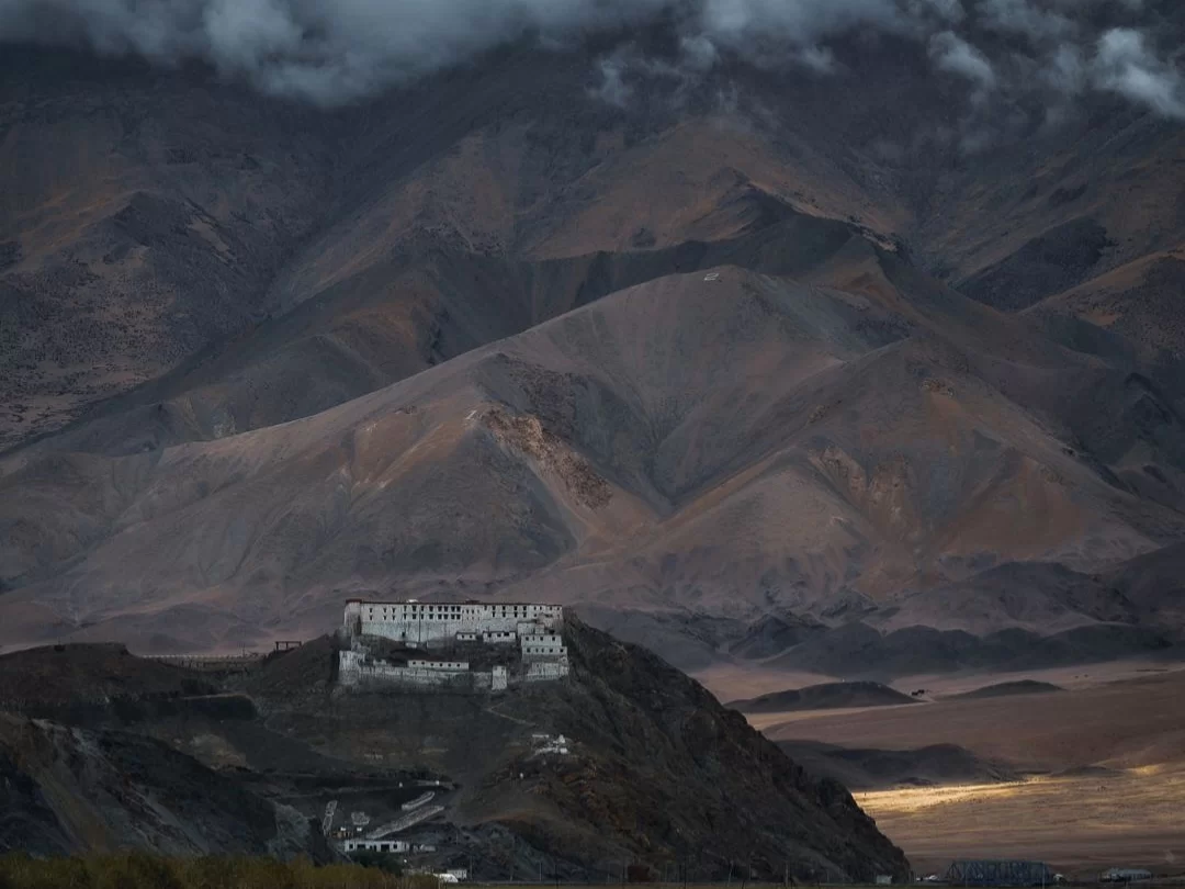 Hanle Gompa perched on hill amid dramatic Ladakh mountains during cloudy dusk, featuring white buildings against barren slopes, perfect Ladakh tour package.