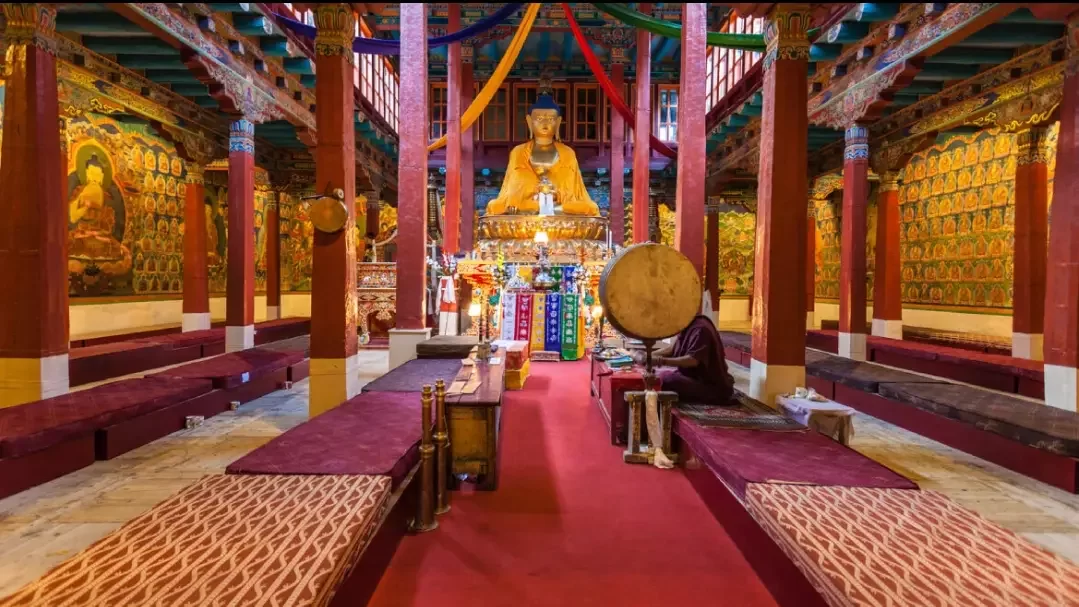 Interior of Dukhang main prayer hall Hemis Gompa Himis Monastery Ladakh India featuring large golden Shakyamuni Buddha statue colorful wall murals red pillars maroon benches monk playing drum prayer flags hanging, serene Tibetan Buddhist gompa ambiance
