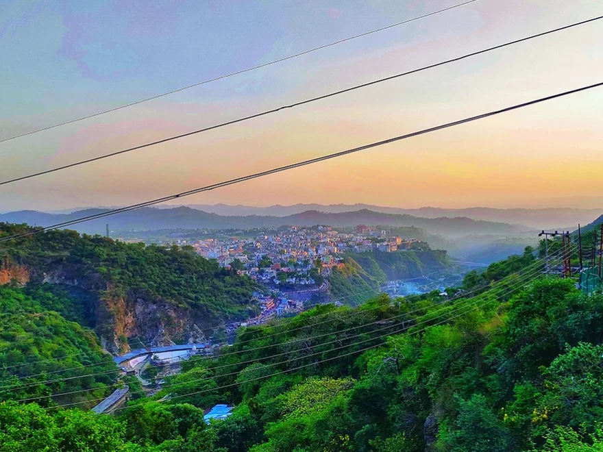 Panoramic view Katra town Jammu Kashmir during golden hour sunset, featuring buildings nestled Trikuta mountains green hills mist power lines valley backdrop Vaishno Devi base, perfect pilgrimage experience Jammu Kashmir tour packages.