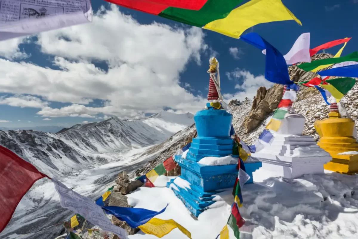 Colorful prayer flags fluttering around blue stupa at Khardungla Pass Ladakh during winter, featuring white yellow stupas snowy mountains cloudy blue sky, perfect adventure experience Ladakh winter road trip package.
