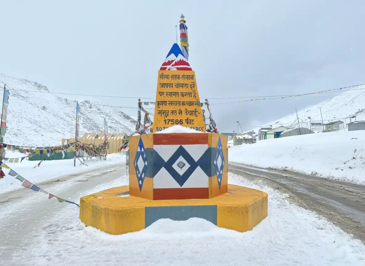 Vibrant yellow chorten prayer flags snowy road at Khardungla Pass Ladakh during winter, featuring Hindi inscription snowy mountains grey sky, perfect adventure experience Ladakh winter road trip package. 