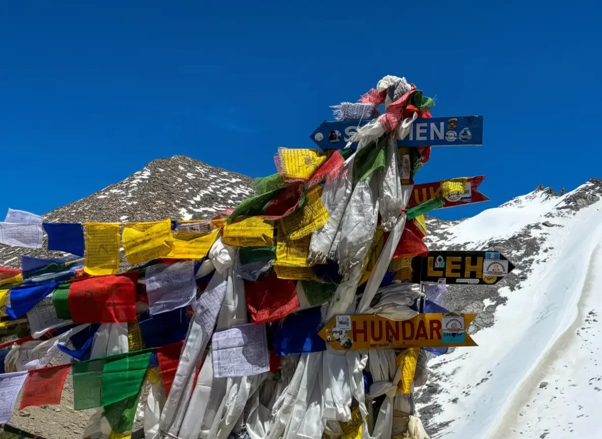 Prayer flags and signposts at Khardungla Pass Ladakh during winter, featuring Nubra Nubra Leh Hunder signs snowy mountains blue sky, perfect adventure experience Ladakh winter road trip package.