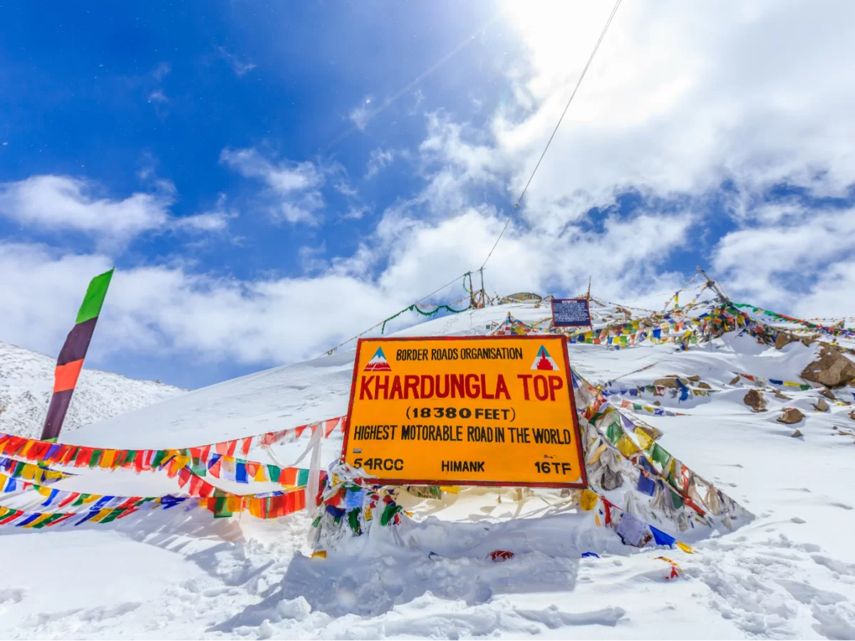 Bright yellow BRO board at snow-covered Khardungla Top (18,380 ft), surrounded by colorful prayer flags under a vivid blue sky, marking one of the world’s highest motorable passes.