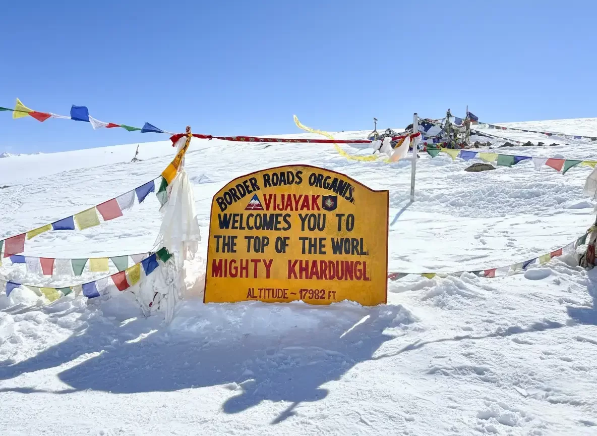 Iconic BRO signboard welcoming to top of world Khardungla at Khardungla Pass Ladakh during winter, featuring prayer flags snowy hill blue sky altitude marker, perfect adventure experience Ladakh winter road trip package.