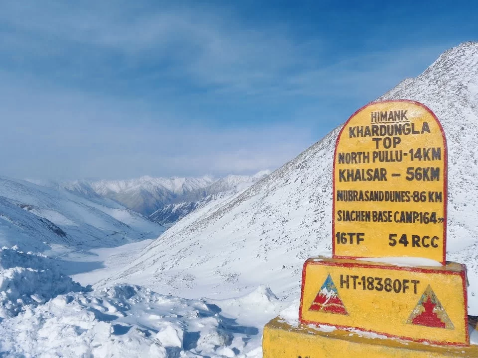 Snowy panorama from Khardungla Top with yellow milestone showing distances to North Pullu, Khalsar, Nubra Sand Dunes, and Siachen Base Camp against rugged Himalayan valleys and
