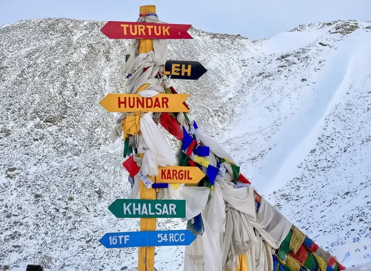 Colorful multi-directional signpost at Khardungla pointing towards Turtuk, Leh, Hundar, Kargil and Khalsar, wrapped in prayer flags against a rugged snow-dusted Himalayan slope.