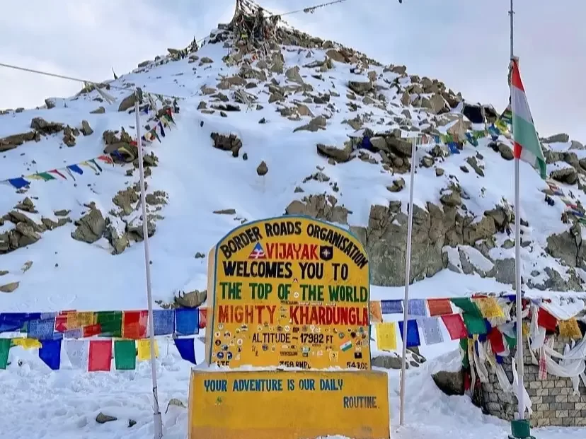 Border Roads Organisation welcome board at snow-clad Khardungla, reading Top of the World Mighty Khardungla, surrounded by prayer flags and rocky Himalayan slopes in the background.