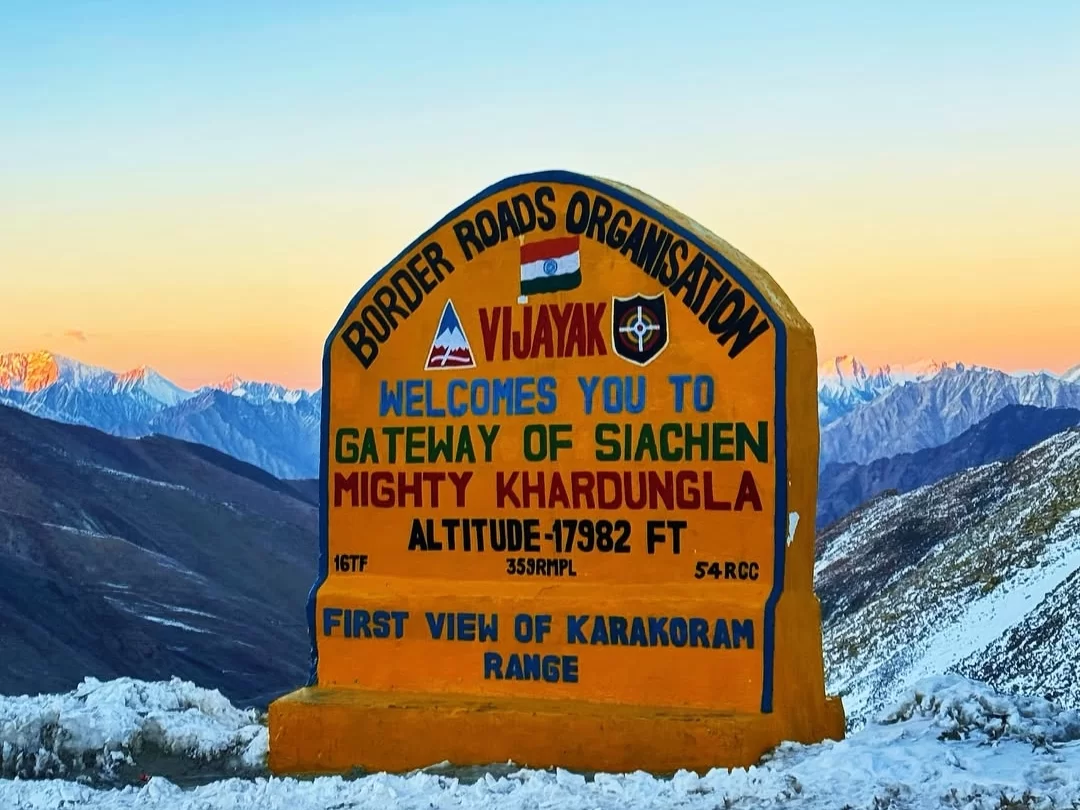 Golden BRO milestone at Khardungla reading “Gateway of Siachen Mighty Khardungla, Altitude 17,982 ft, First View of Karakoram Range,” set against snow-dusted mountains at sunrise or sunset.