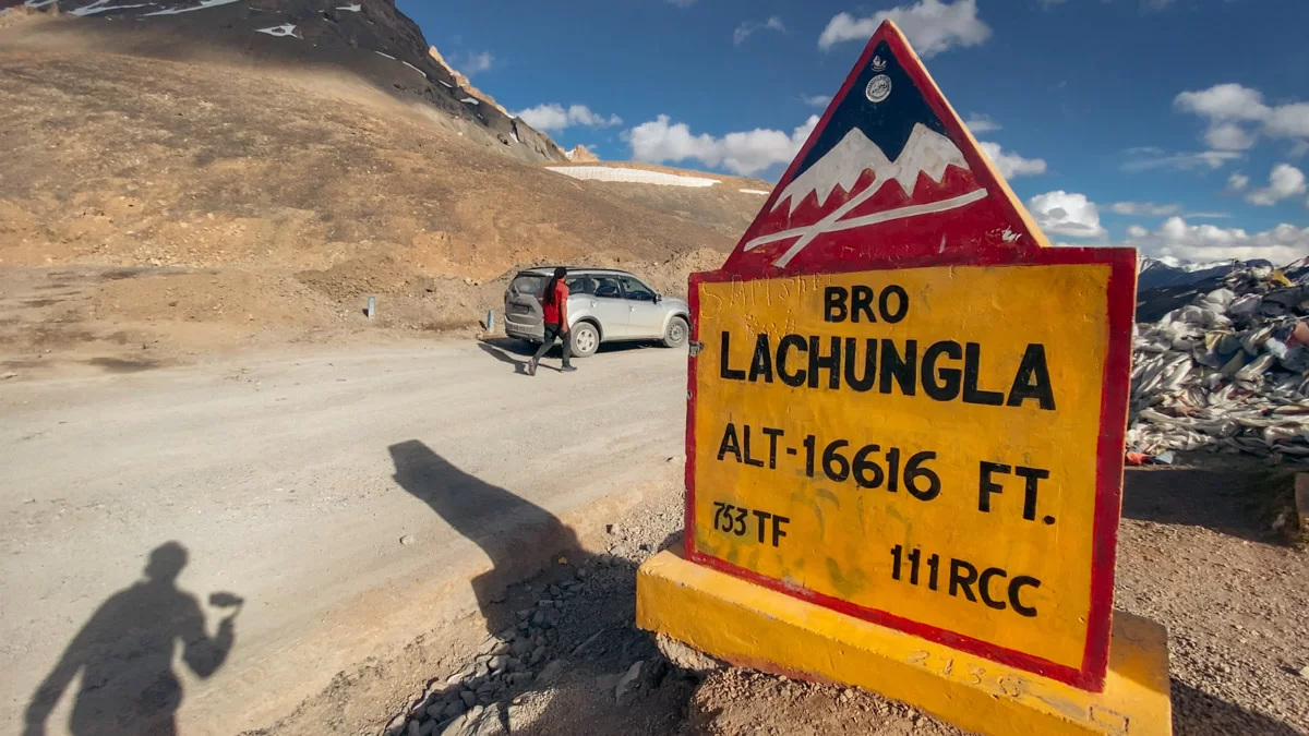 Lachulung La Pass BRO sign at 16,616 ft Leh-Manali Highway in clear weather, featuring mountains, parked car and tourist, perfect adventure Ladakh tour package.