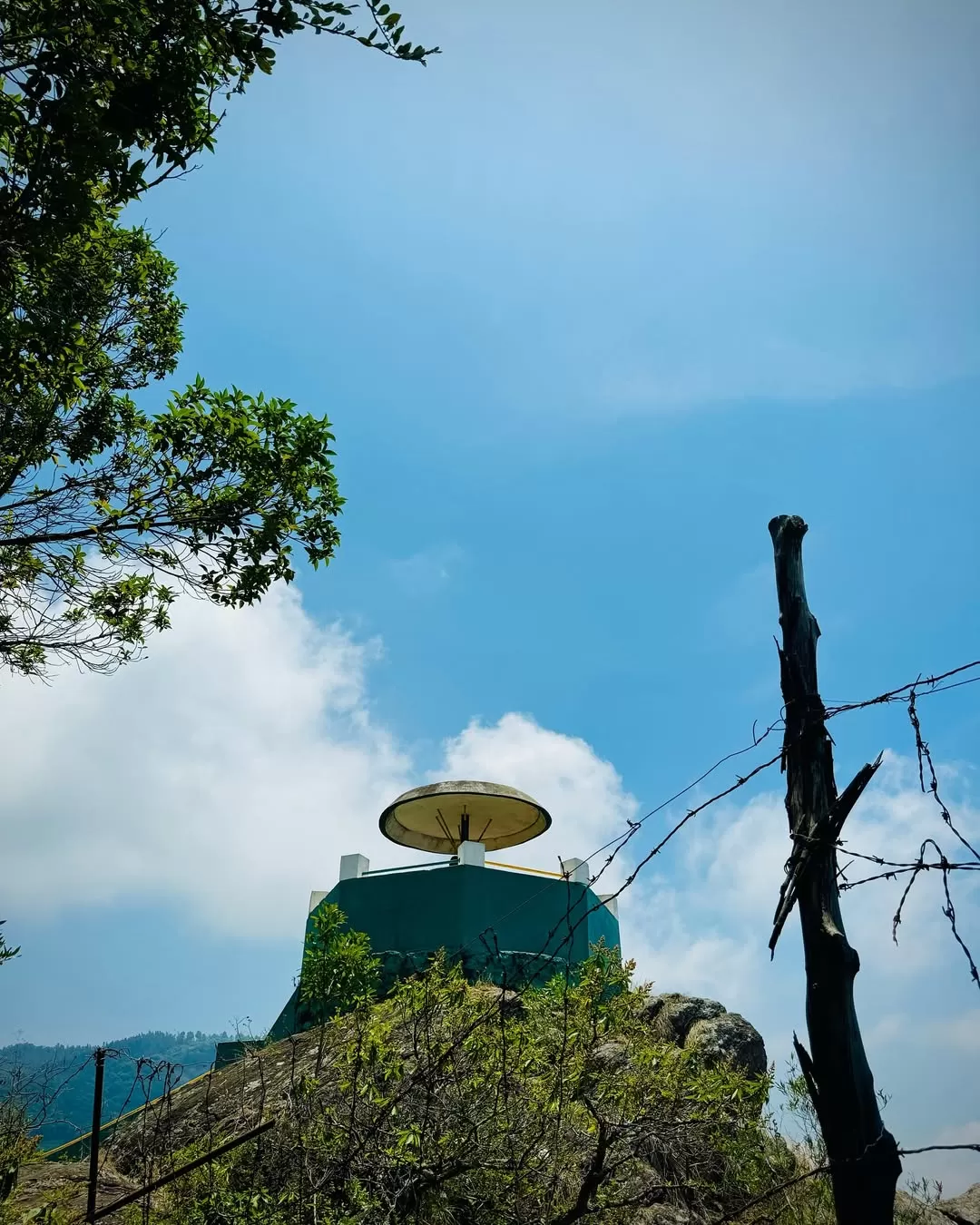 Raja's Seat viewpoint at Madikeri Coorg during sunny day, featuring green tower with yellow umbrella roof, trees, rocks, perfect panoramic experience Karnataka tour packages.